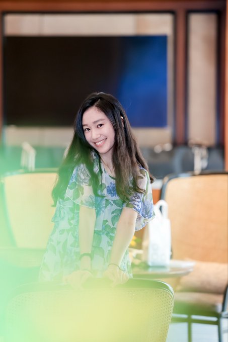 A young woman with long dark hair smiles warmly while leaning on a chair in what appears to be a lounge or lobby area, with a soft green/yellow light leak effect in the foreground.