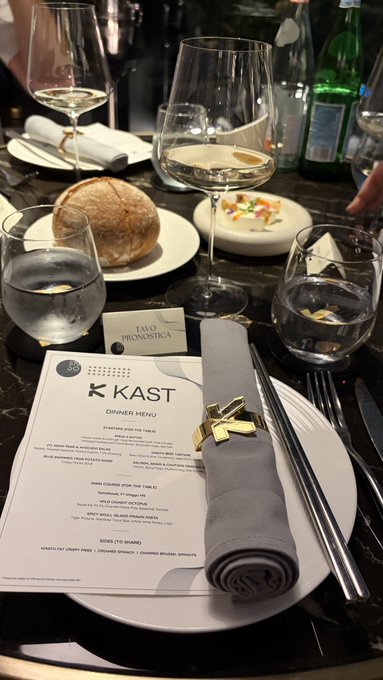 A formal restaurant table is set with wine glasses, water glasses, bread, a small dessert or appetizer, cutlery, and a gray rolled napkin secured with a gold bow. The visible menu reads “KAST DINNER MENU,” with cards labeled “FAVA PROSCIUTTA” and another partially obscured item, matching the post’s tag for @KASTxyz.