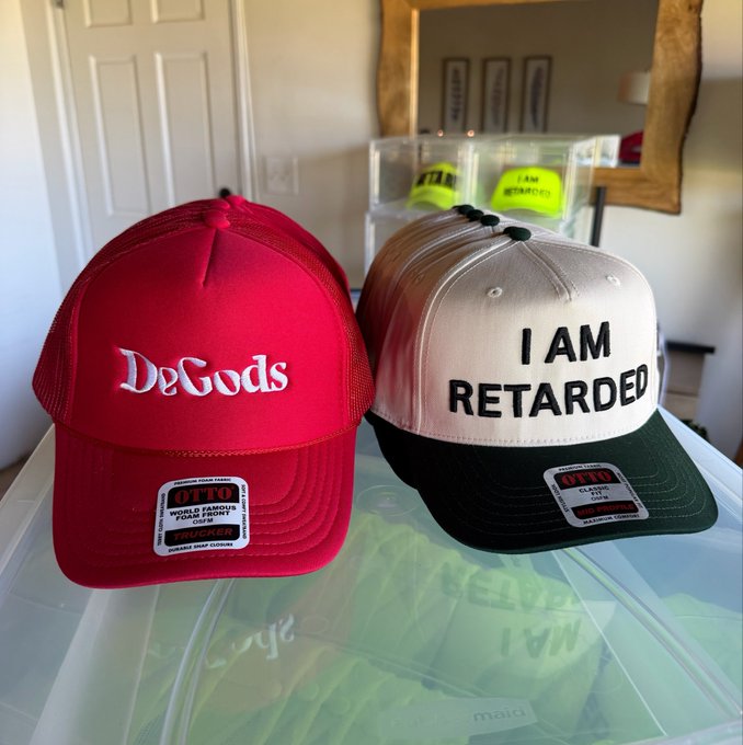Two baseball caps sit on a surface: a red OTTO 'World Famous' cap with 'DeGods' embroidered, and a cream/dark green OTTO snapback cap with 'I AM RETARDED' embroidered.