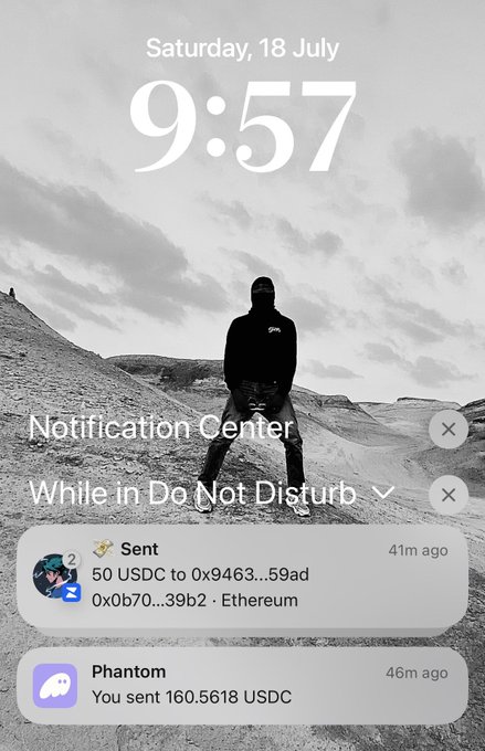iPhone lock screen displaying Saturday, 18 July at 9:57 with notifications including a Phantom wallet confirmation that 160.5618 USDC was sent and another 50 USDC sent to an Ethereum address.