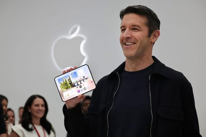 A man with short dark hair wearing a dark jacket over a navy shirt is smiling while holding up a foldable smartphone displaying its home screen, with an illuminated Apple logo visible on the wall behind him.
