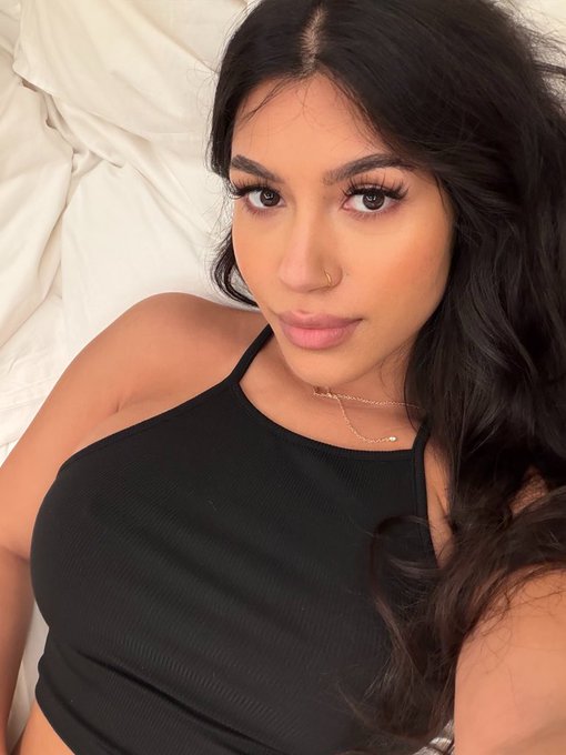A young woman with long dark hair and a nose ring takes a selfie while wearing a black halter top, lying against white bedding.
