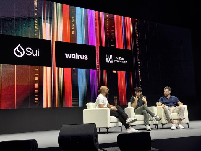 A panel of three people sit in white armchairs on stage at a tech event, with large screens behind them displaying logos for "Sui," "walrus," and "The Data Foundation" against a vibrant, multicolored striped backdrop. The image illustrates the breaking news that The Data Foundation, formerly known as Story Protocol, is retiring its Layer 1 blockchain to operate natively on the Sui network, visually connecting the two brands at the center of this strategic shift.