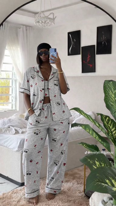The image features a woman wearing a pajama set with red hearts on her pants. She is standing in front of a bed that has white sheets and a green plant nearby, possibly a houseplant or a decorative item. The woman appears to be taking a selfie using her cell phone while posing for the camera.