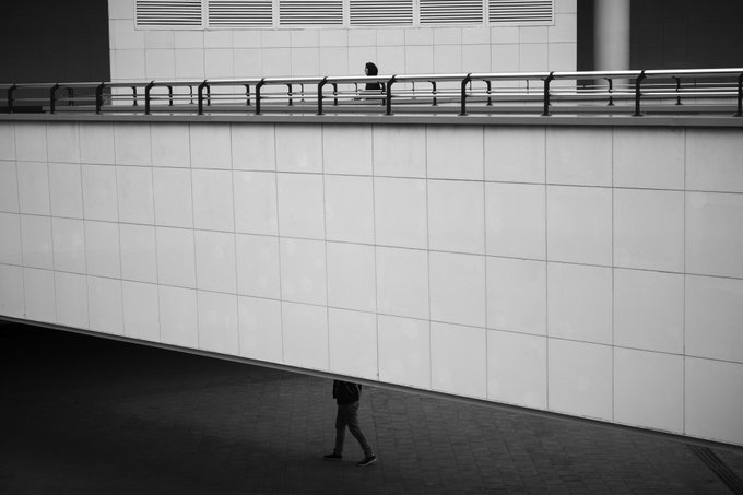 A black and white photograph shows a minimalist architectural scene with a person walking on an elevated walkway above another person walking on the ground level below, creating a stark geometric composition.