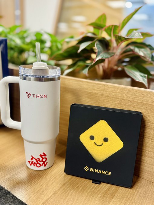 A white travel mug branded with the 'TRON' logo next to a black box featuring a yellow smiley face and the 'BINANCE' logo, displayed on a wooden surface.