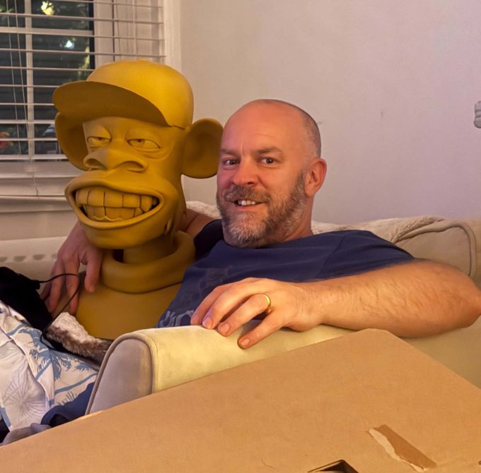 A bald, bearded man wearing a dark t-shirt sits on a couch, smiling at the camera while his arm rests around a large, grinning yellow monkey statue wearing a cap.
