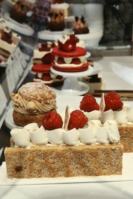 A display case at a bakery showing an assortment of elegant desserts, including a long rectangular almond cake topped with cream rosettes, fresh raspberries, and white chocolate shards.