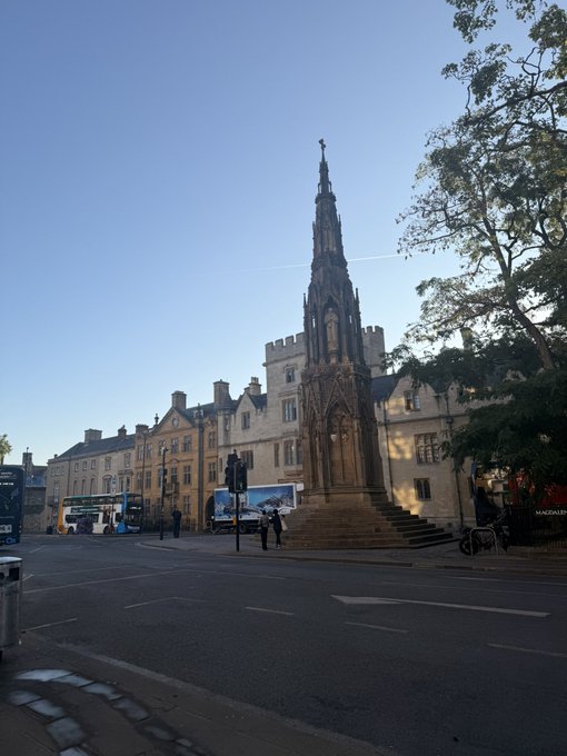 A photograph of a tall, gothic-style stone monument in a town square, with buses and historic buildings in the background under a clear blue sky.