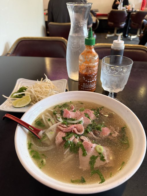 A bowl of Vietnamese pho with rare beef slices, onions, and cilantro sits on a dark table, accompanied by a side plate of bean sprouts and lime, a bottle of sriracha sauce, and a glass of ice water.