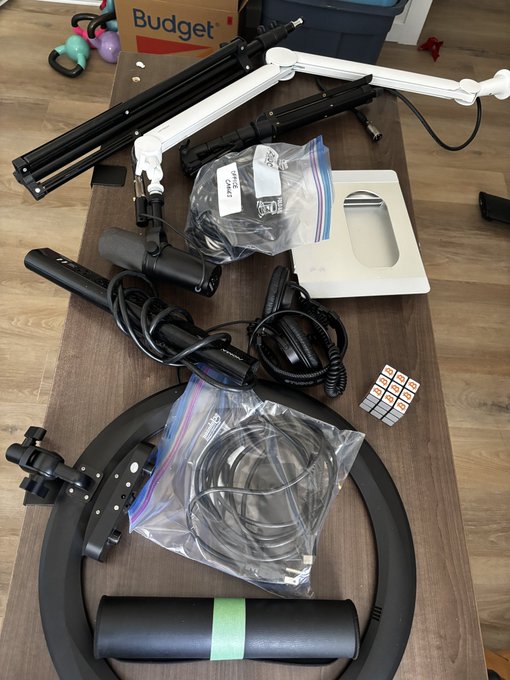 A top-down view of various microphone stands, boom arms, cables, and ring light components laid out on a wooden table.