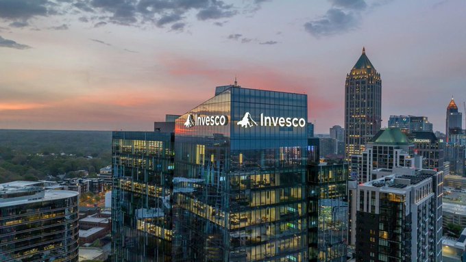 A glass skyscraper prominently displays the "Invesco" logo across its facade at dusk, with Atlanta's skyline and a pink-hued sunset in the background. The image showcases Invesco's headquarters as the asset management giant moves to enter the stablecoin market.