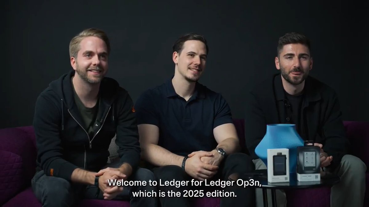 Three men sit on a purple couch during a Ledger Op3n panel, with a blue speaker and hardware wallet devices displayed on a table beside them, while on-screen text reads: "Welcome to Ledger for Ledger Op3n, which is the 2025 edition."