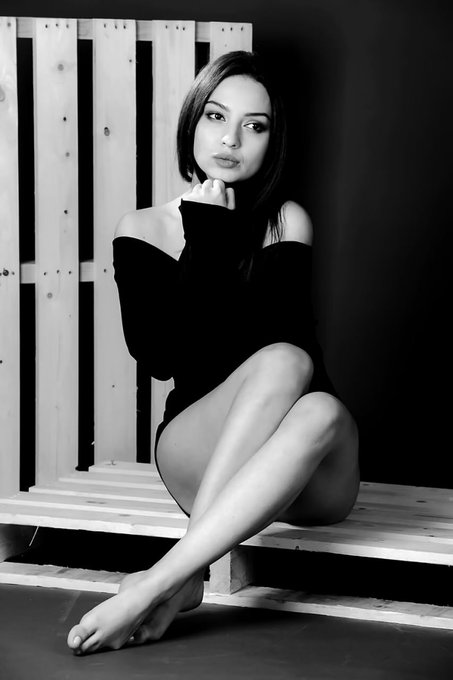 Black and white portrait of a young woman with long dark hair, wearing a black off-shoulder top, sitting on a wooden pallet against a slatted wall.