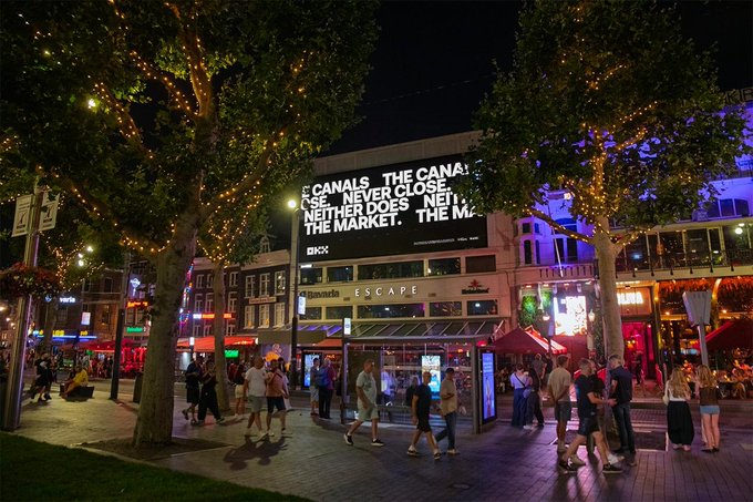 A nighttime urban plaza scene with string lights illuminating trees, pedestrians walking, and a large illuminated billboard reading 'THE CANALS NEVER CLOSE. NEITHER DOES THE MARKET. THE M...' above a 'CANAL ESCAPE' storefront.