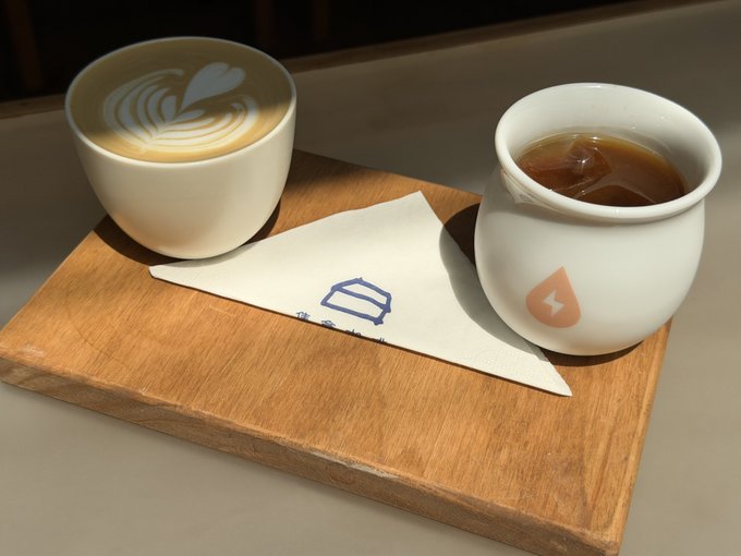 Two beverages—a latte with a heart-shaped latte art and an iced coffee—served on a wooden tray with a napkin printed with a blue cup logo.