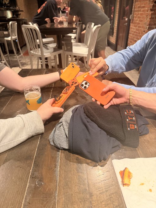 Two people at a wooden table examining orange Nintendo Switch cases and an orange phone case, with a brown beanie and a plate with a slice of pizza nearby.