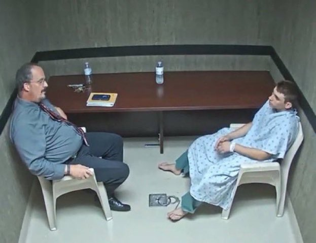 A photograph of two men sitting in plastic chairs across from each other at a table in a sparse room resembling an interrogation room, with one man in a suit and tie and the other in a hospital gown.