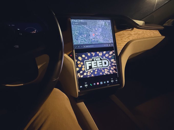 A Tesla vehicle's center console touchscreen displays a navigation map and 'THE FEED' app interface with colorful graphics.