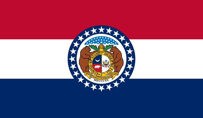 The Missouri state flag, featuring its red, white, and blue stripes with the central seal displaying two bears, a shield, and the date "MDCCCXX" (1820), is shown as the backdrop to the news about three Missouri men charged in an attempted Bitcoin robbery.