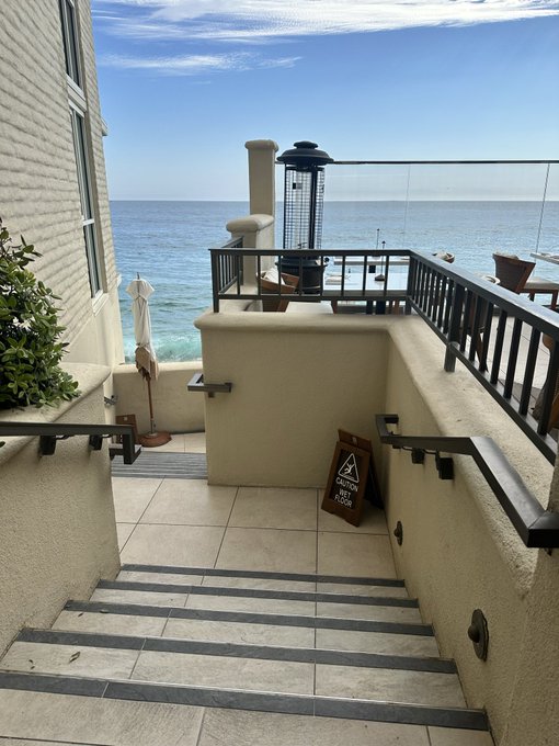 An oceanfront patio with tile steps leading down, a patio heater, and a view of the blue sea under a partly cloudy sky, with a 'CAUTION WET FLOOR' sign visible.