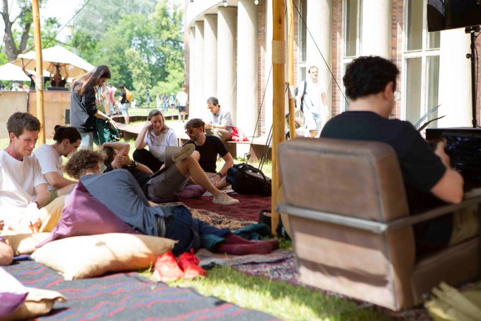 People relax on colorful pillows and rugs on a sunny outdoor patio, with some sitting casually on the ground chatting while others walk by in the background.