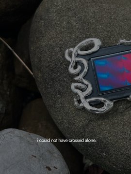 A metallic snake ornament coils around a screen displaying vibrant red and blue hues, resting on a stone. The caption reads "i could not have crossed alone," aligning with the "æon" project's evocative and atmospheric theme.