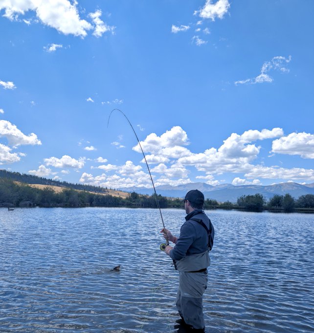 An angler stands waist-deep in a serene lake, casting a fly rod toward the mountains under a bright, cloud-dappled sky—embodying the post's call to "touch grass" by trading screens for fresh air and the rhythm of the water.