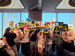 A group of people posing together, their faces obscured by various emoji stickers, in what appears to be an office or modern interior space.