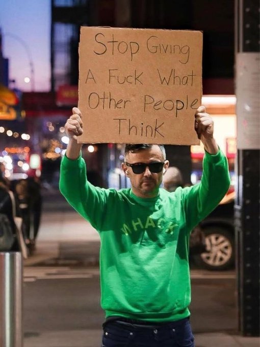 A man wearing a green sweatshirt and sunglasses holds up a cardboard sign on a city street that reads "Stop Giving A Fuck What Other People Think".