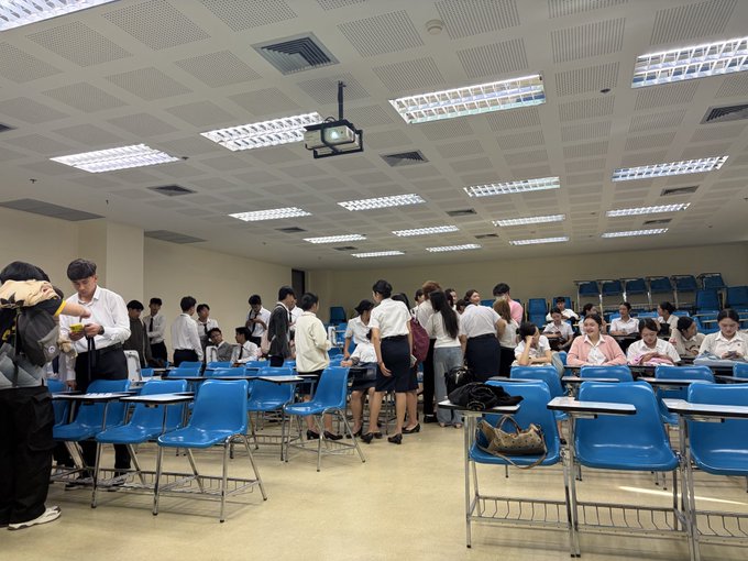 A crowded university classroom or lecture hall with blue chairs and desks, where students are standing and interacting, likely during a break.