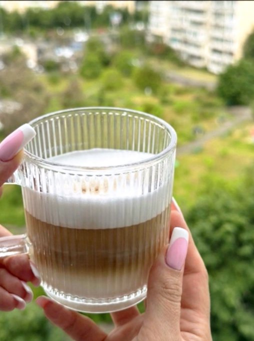 The image features a person holding a glass mug filled with a latte, which appears to be freshly made. The person's hands are positioned over the cup, showcasing its contents. In the background, there is a cityscape visible through an open window, providing a serene view of urban life outside. The post about oil and stocks also includes a link to another website, possibly related to the coffee industry or financial markets.