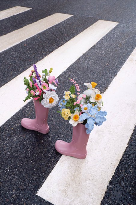 Two pink rubber rain boots repurposed as decorative flower vases filled with colorful artificial flowers, placed on a black and white crosswalk.