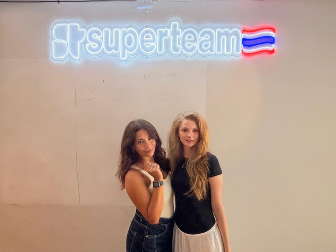 Two young women pose beneath a glowing "superteam" neon sign with a stylized logo, smiling for the camera in what appears to be a casual, welcoming atmosphere—fitting the cheerful "GM from Superteam Thailand" greeting.