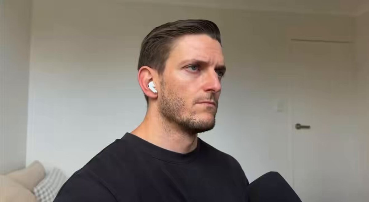 The image features a man wearing a black shirt sitting down, possibly watching TV or using his phone. The background shows a white wall, which contrasts with the man's dark clothing. There are two other people visible in the image, but they are not the main focus of the scene.