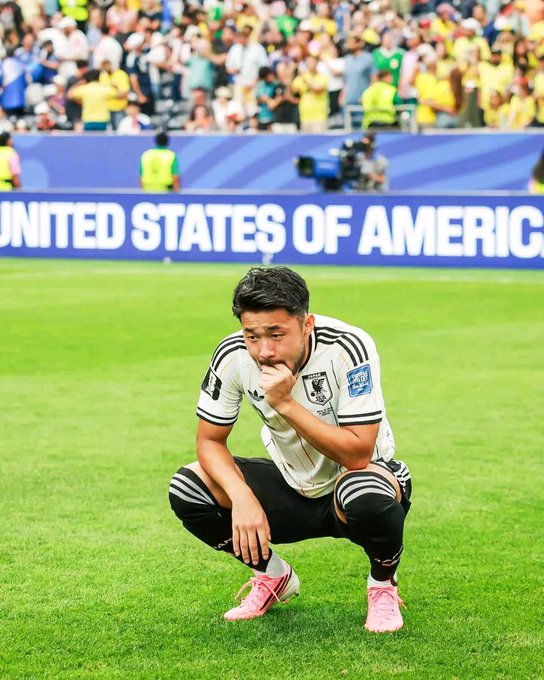 Photograph of a German national football team player in a white kit crouching on the pitch in a thoughtful pose, with a stadium crowd and 'UNITED STATES OF AMERICA' banner in the background.
