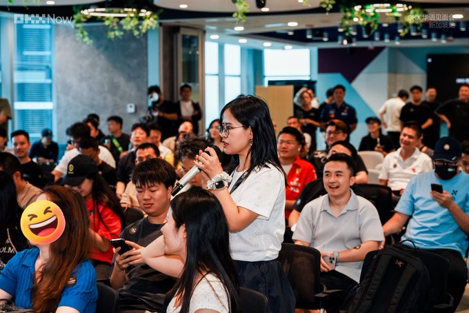 A woman speaks into a microphone during a conference or event, with an attentive audience seated behind her.