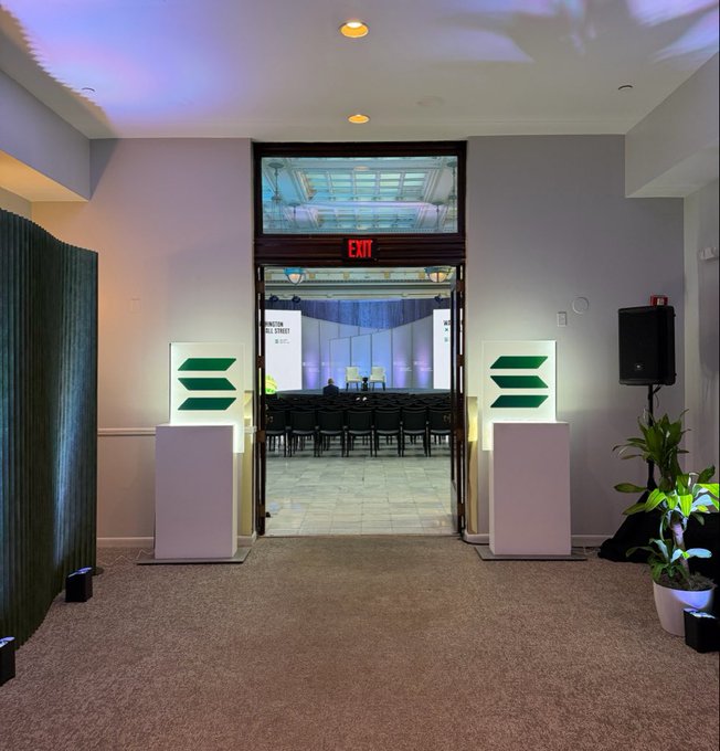 A view through an open doorway with an illuminated EXIT sign above it, flanked by two white pillars displaying green chevron signs, leading into a hall with rows of chairs and a stage lit in blue.