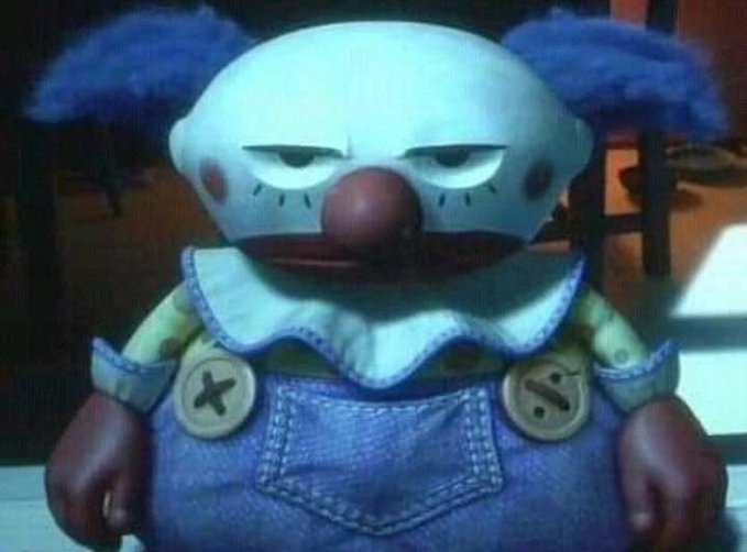 A close-up of a creepy clown doll with blue fuzzy hair, a white face, a red nose, and blue overalls with large buttons.