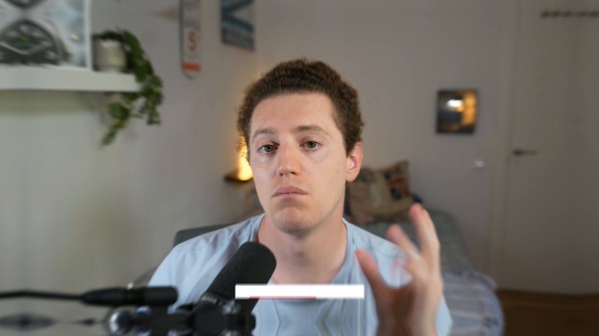 The image shows a man sitting at a desk with a microphone in front of him. He appears to be making a hand gesture, possibly giving instructions or explaining something related to cryptocurrency. The background features a white wall with a clock hanging on it, indicating that this scene takes place indoors during the day.