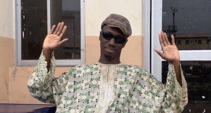 A man in a green floral patterned tunic, flat cap, and sunglasses poses with both hands raised in front of an orange building.