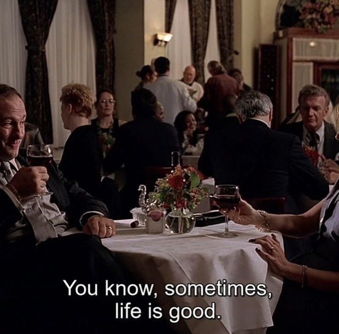 A man and woman sit at a white-clothed restaurant table with wine glasses, surrounded by other diners in a dimly lit, elegant setting, with the caption 'You know, sometimes, life is good.'