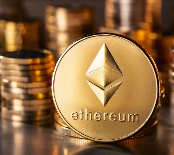 A close-up photograph of a gold coin featuring the Ethereum logo and the word 'ethereum', with stacks of additional coins blurred in the background.