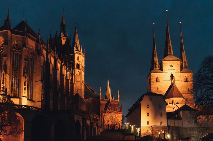 A nighttime photograph of illuminated Gothic cathedral spires and towers against a deep blue sky, likely Erfurt Cathedral in Germany.