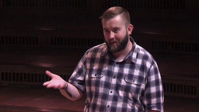 A bearded man in a plaid shirt gestures with his hand while speaking on a stage with a dark wooden backdrop.