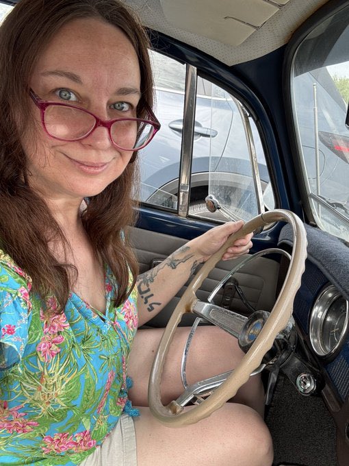 A woman with red-framed glasses and a tattoo reading "sol" smiles while sitting behind the steering wheel of a vintage car. The image's tattoo and casual setting align with the post celebrating her cohosting of a space themed around $SOL.