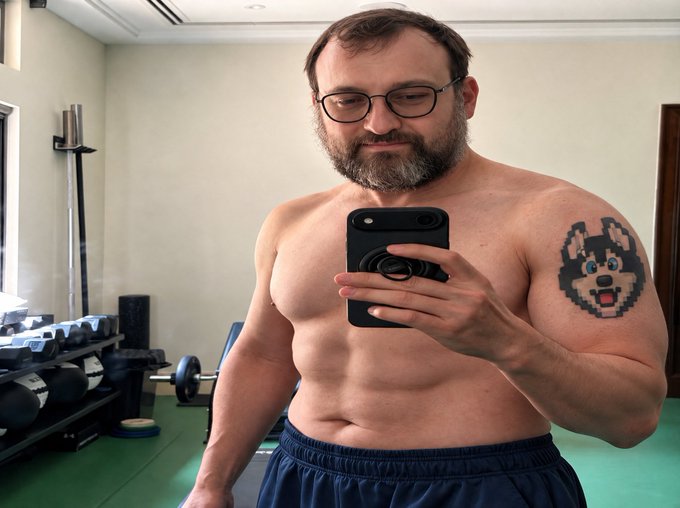 A shirtless, muscular man with glasses and a beard taking a mirror selfie in a gym, displaying a pixelated dog tattoo on his left shoulder.