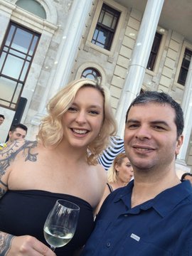 The image features two people posing together with wine glasses in their hands. One person has blonde hair and tattoos, while the other has dark hair and no visible tattoos. They are both smiling for the camera, creating a friendly atmosphere. The background includes an old building or a large stone structure, which adds to the overall ambiance of the scene.