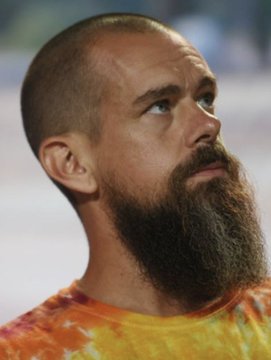A close-up portrait of a man with a long brown beard and shaved head, wearing a yellow and orange tie-dye shirt.