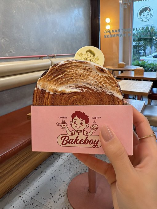 A hand holding a pink card with the 'Bakeboy' logo in front of a large pastry topped with toasted meringue, taken inside a cafe.